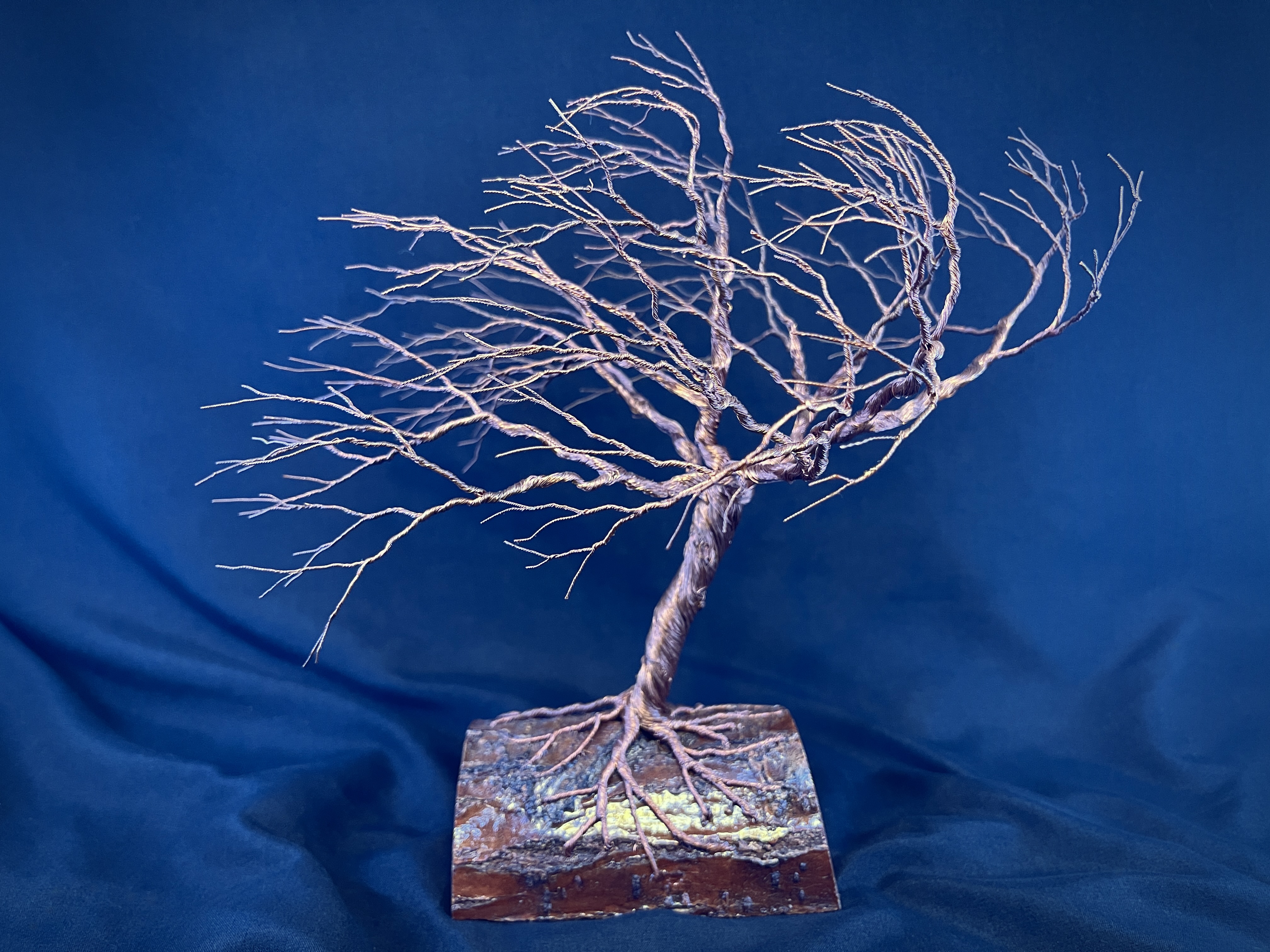 Dramatic windswept copper wire tree rooted on a rectangular base against deep blue