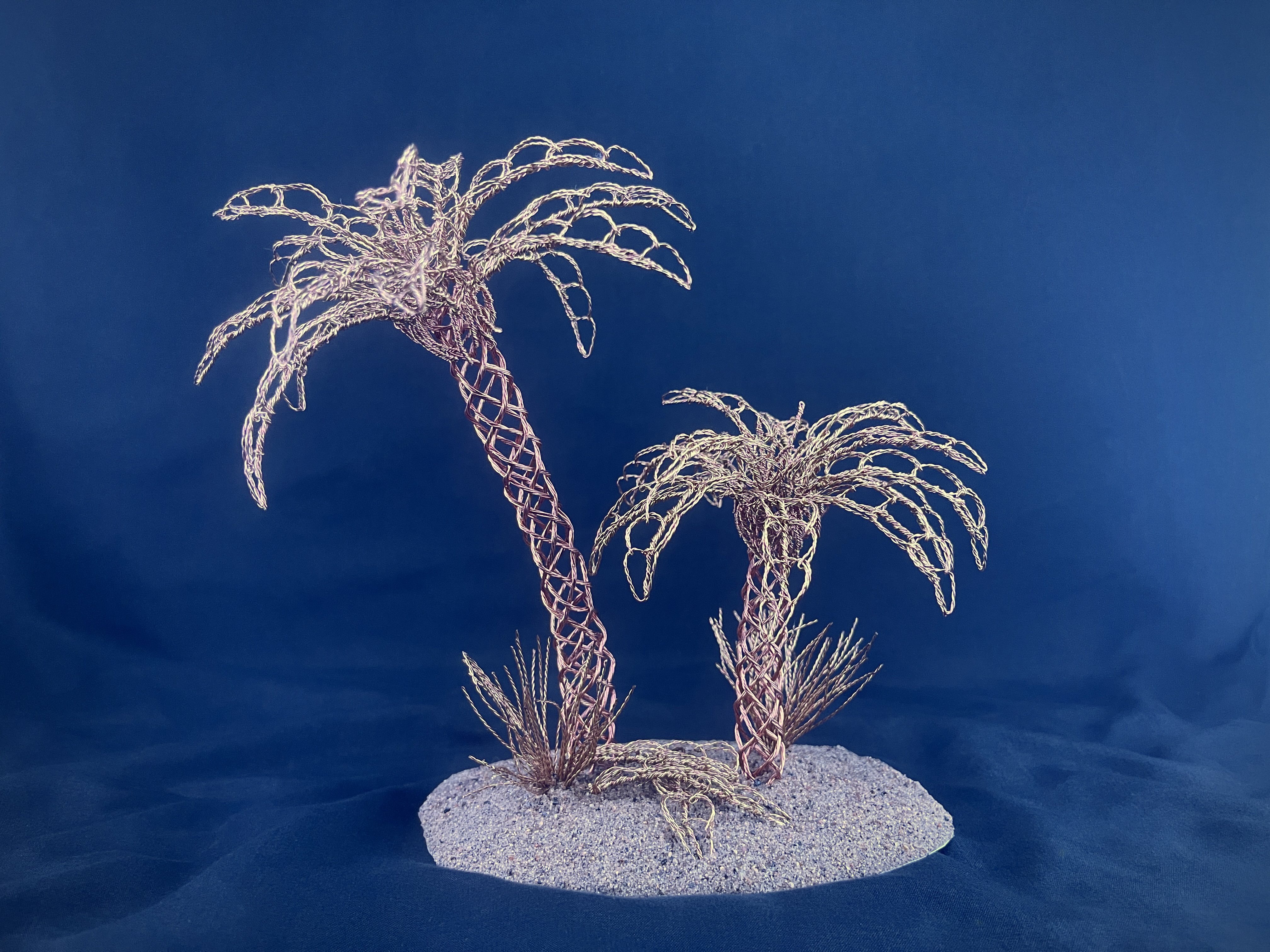 Two golden wire palm trees on a sandy base against blue
