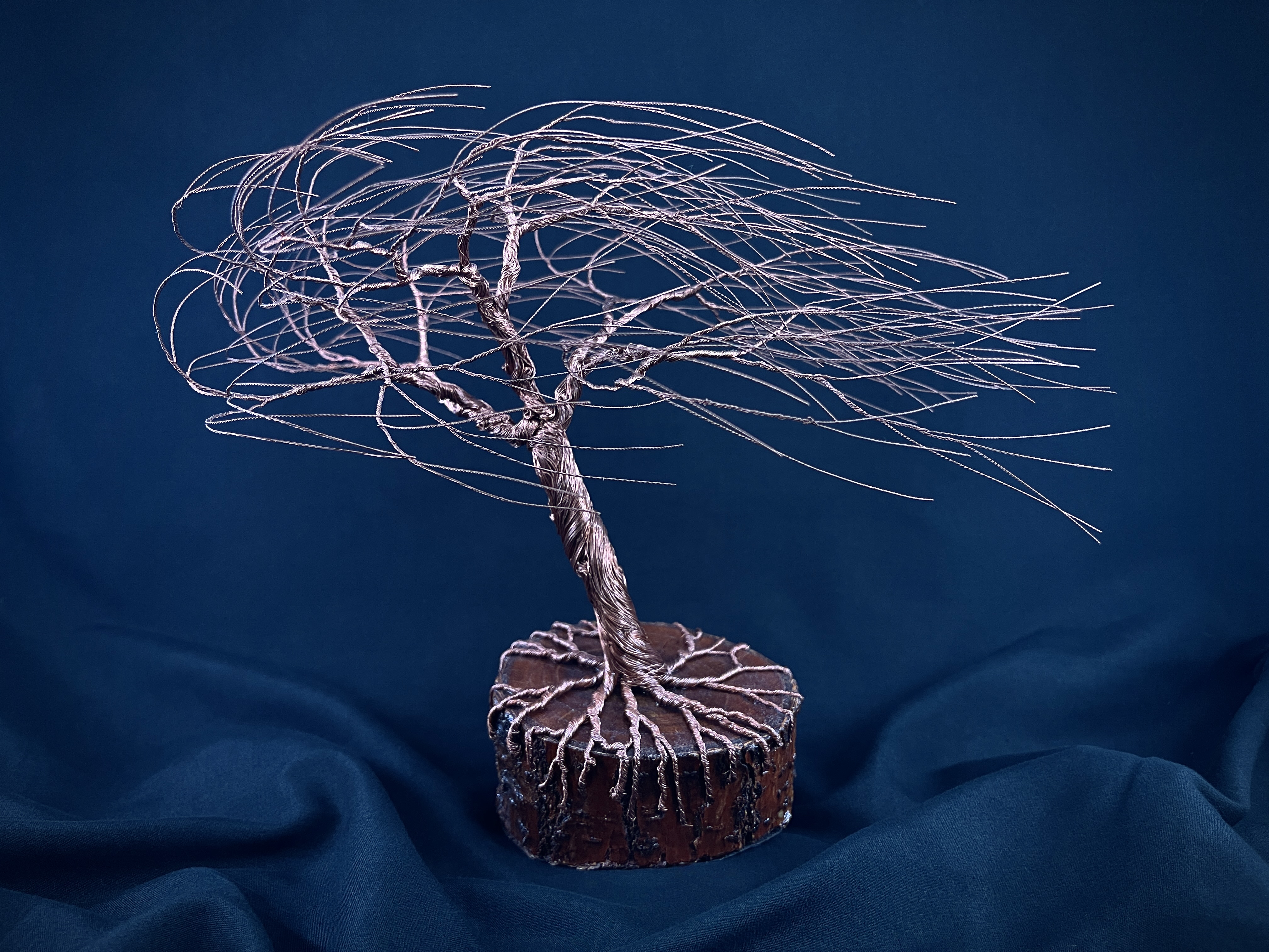 Windswept copper wire tree sculpture rooted on a round wood base against deep blue